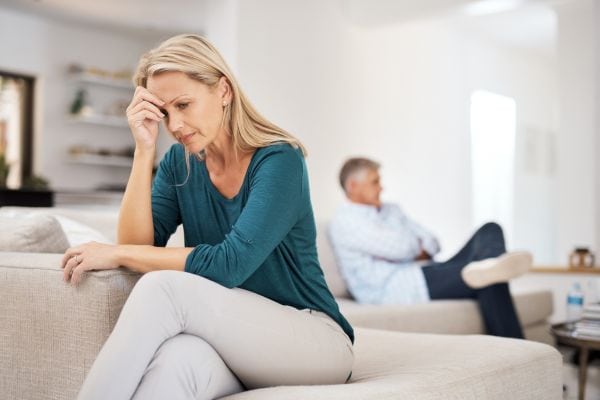 Eine nachdenkliche Frau sitzt im Vordergrund auf einem Sofa, während ein Mann mit Abstand hinter ihr sitzt und zur Seite blickt.