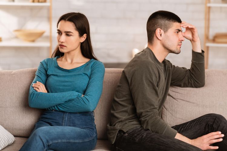 Ein Paar sitzt Rücken an Rücken auf einem Sofa. Die Frau hat die Arme verschränkt und blickt zur Seite, während der Mann sich mit einer Hand an die Stirn fasst.