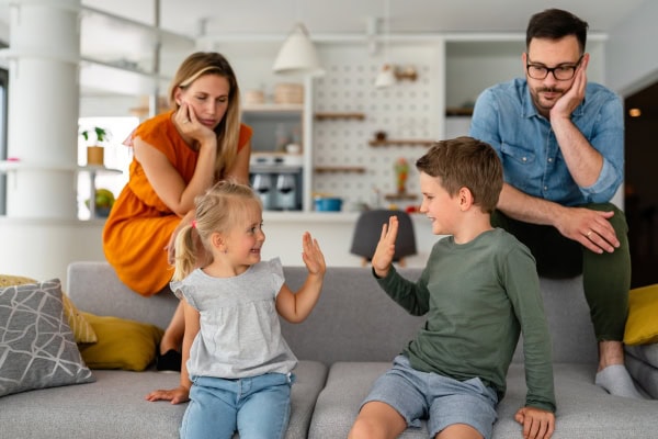 Zwei Kinder spielen auf dem Sofa miteinander, während ihre Eltern im Hintergrund sitzen und erschöpft wirken