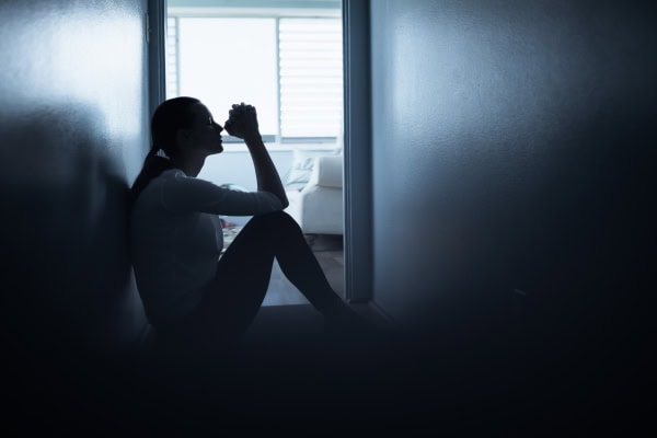 Eine Frau sitzt allein in einem dunklen Flur auf dem Boden und stützt nachdenklich die Hände vor ihr Gesicht.
